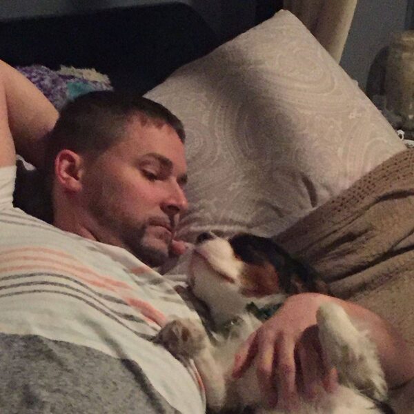 A man asleep on the couch with a Cavalier puppy resting against his arm