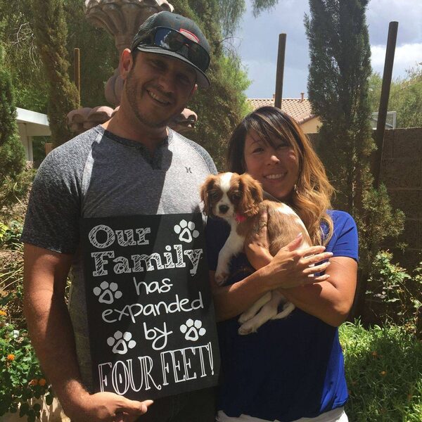 A man and woman holding their new puppy, beside a sign reading Our Family Has Expanded By Four Feet